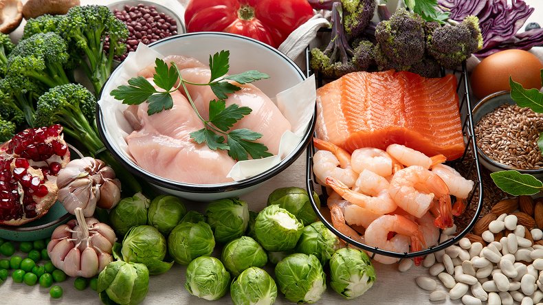 Assorted fresh ingredients including raw fish, shrimp, salmon, vegetables, legumes, and grains arranged on a white surface.